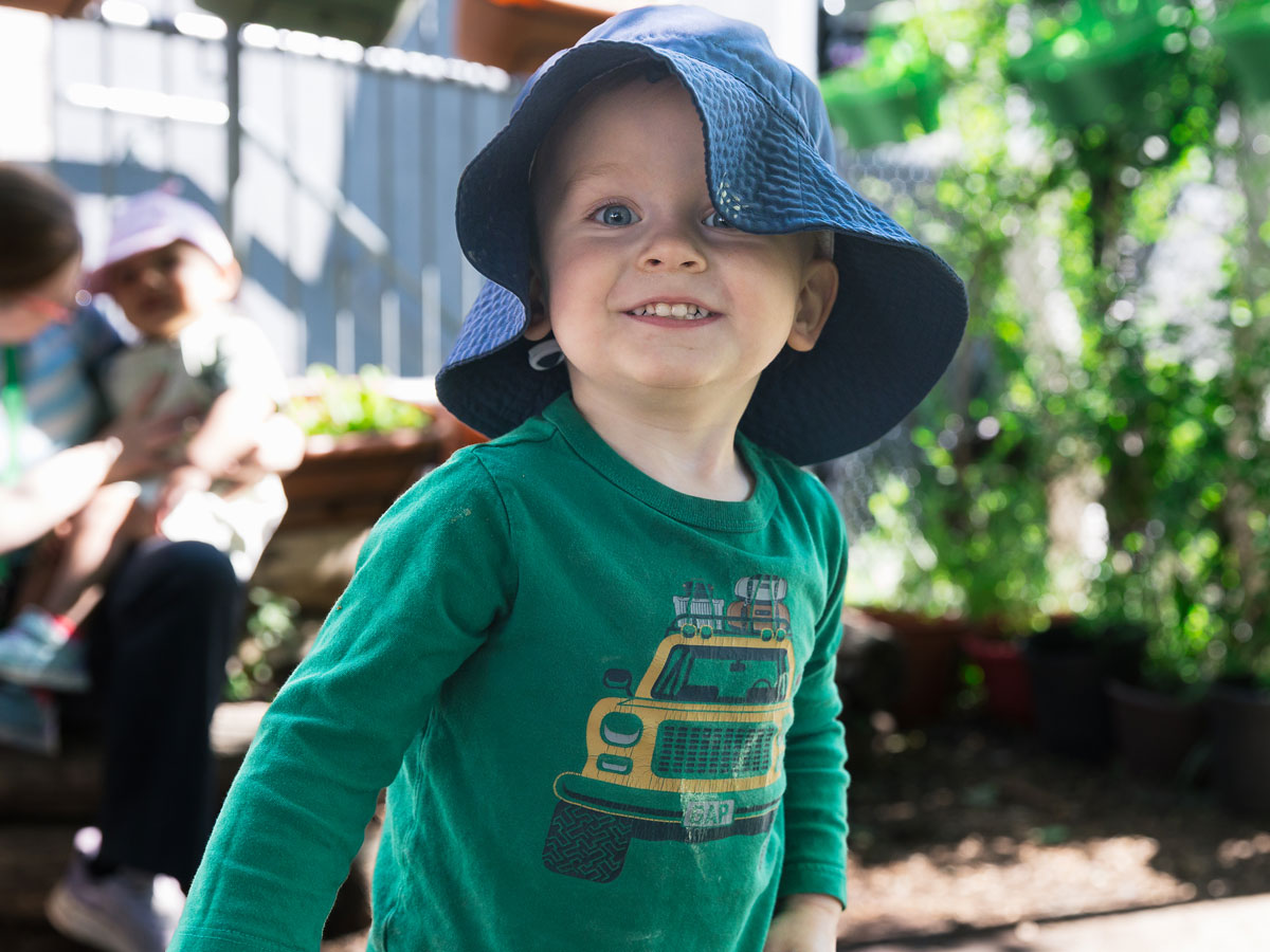 A candid photo of a male toddler inside Green Apple Kids' natural playground, smiling at the camera. A teacher holding another child on her lap can be seen in the background.