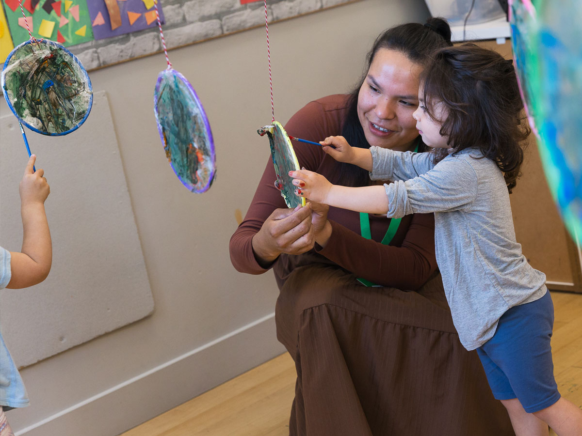A candid photo of a smiling female ECE teacher kneeling beside a preschooler painting a dreamcatcher piece of art hanging from a braided string.