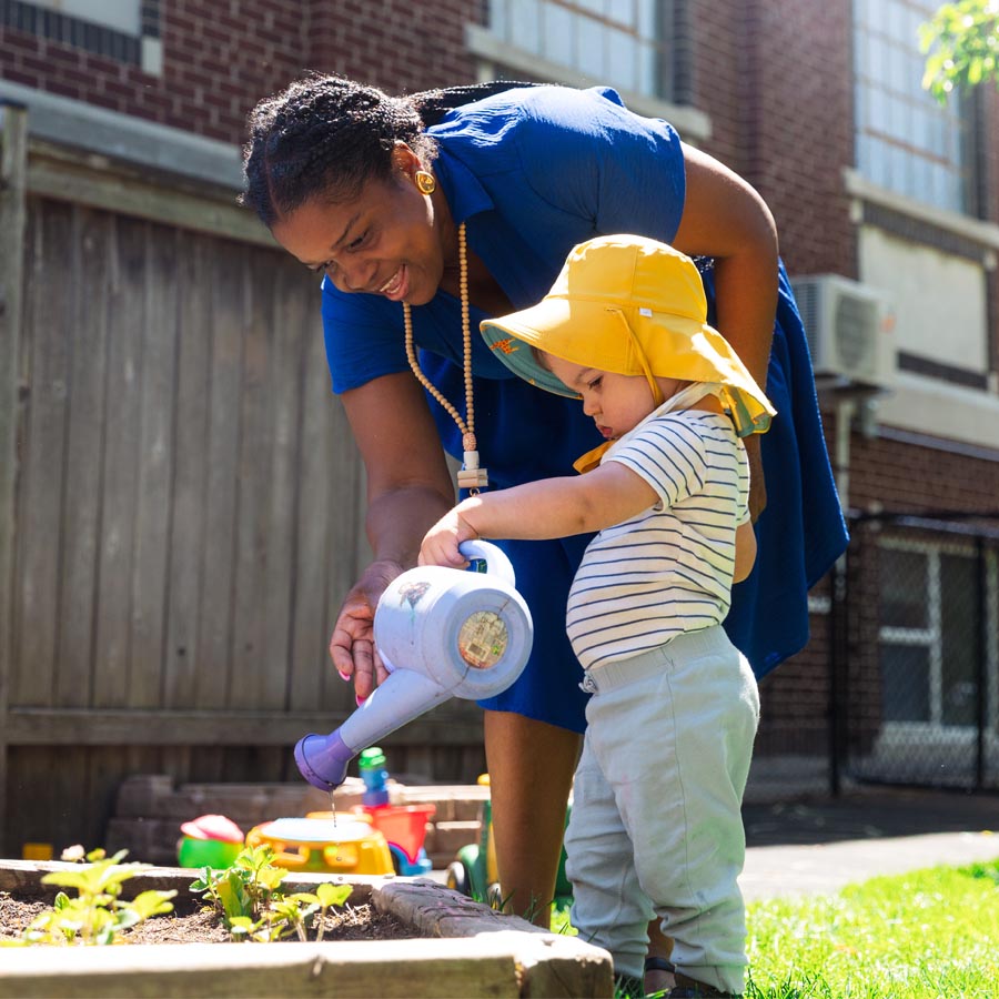Candid photo of a female ECE teacher helping an infant pour water from a watering can onto herbs in a garden bed at Green Apple Kids, a West End Toronto daycare. 