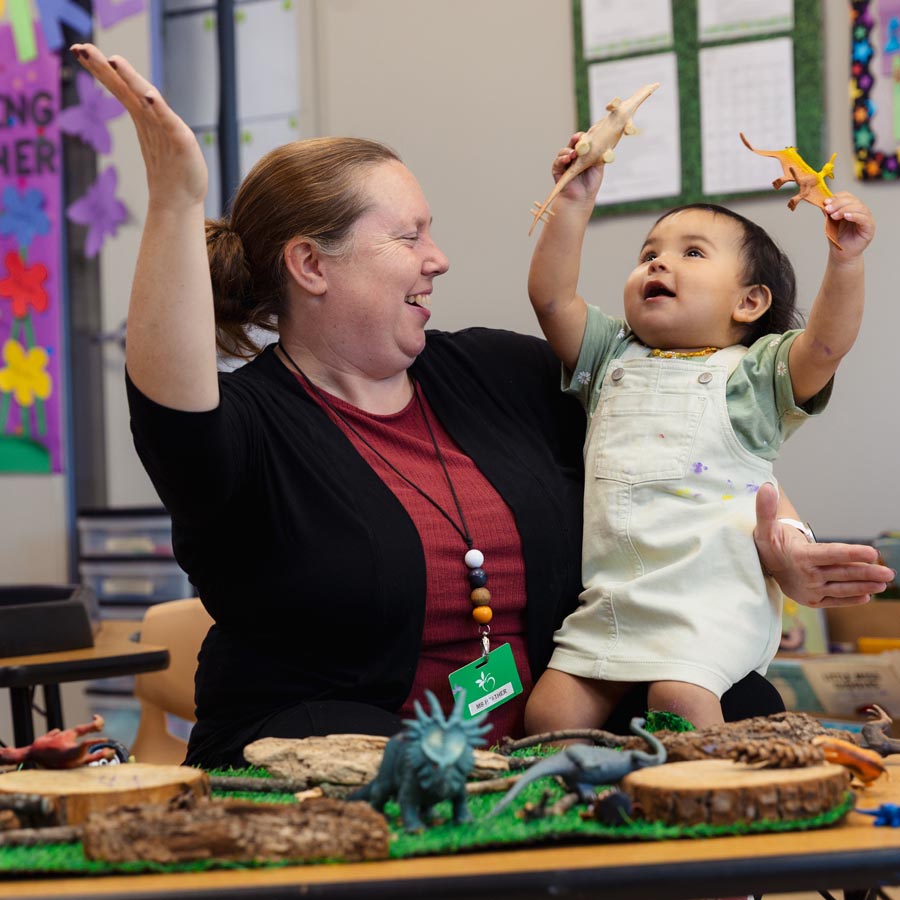 In-class candid photo of a female ECE teacher balancing a toddler on her lap who is holding dinosaur figures up in both hands. Both the teacher and child are smiling.