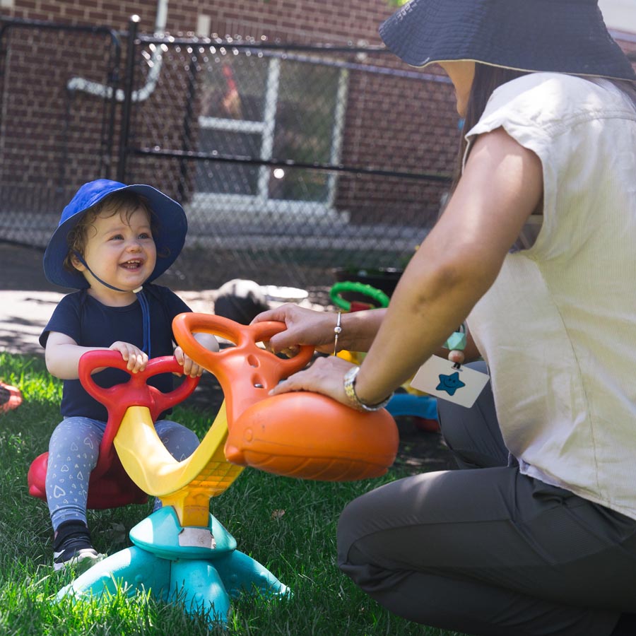 A sunny outdoor candid shot of an infant smiling at the teacher, while the teacher pushes down on a small teeter totter, in Windermere Kids natural playground in Bloor West Village Toronto.