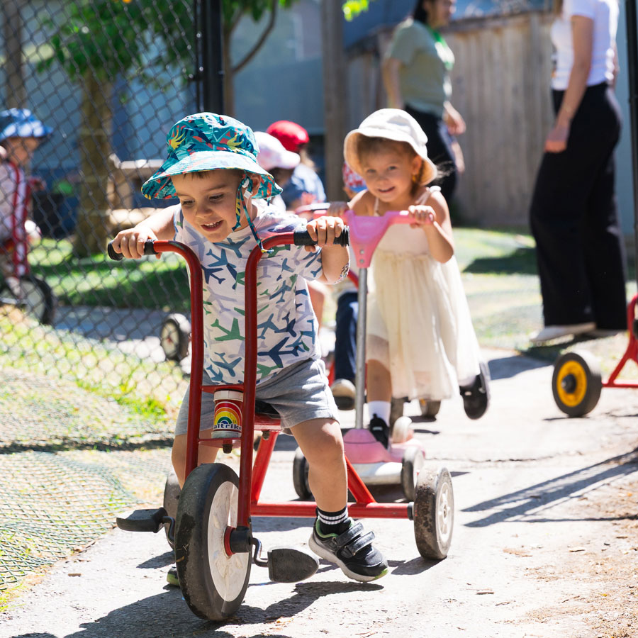A candid photo of two preschool students riding tricycles on the outdoor track in the natural playground at Green Apple Kids, a Toronto West End daycare. Both children are smiling. Other children and teachers are in the background out of focus.