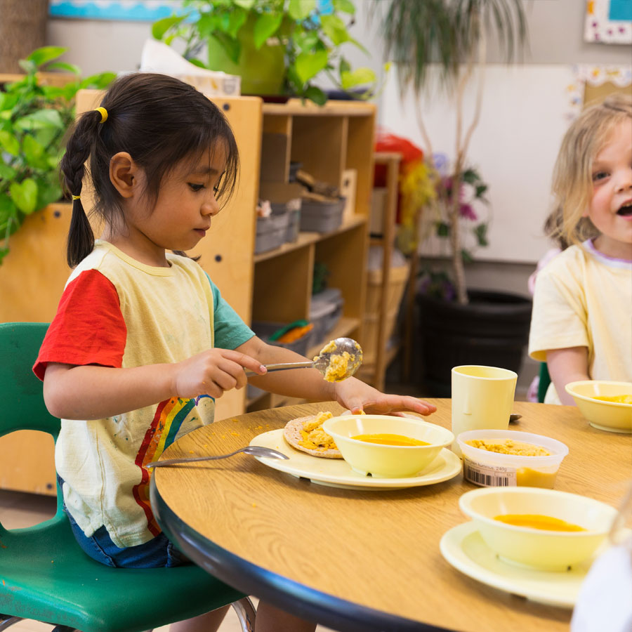 Two female preschool students sitting at a table eating lunch. One student is smiling at the camera while the other student is focused on scooping humus on to a pita with a bowl of soup on the same dish.