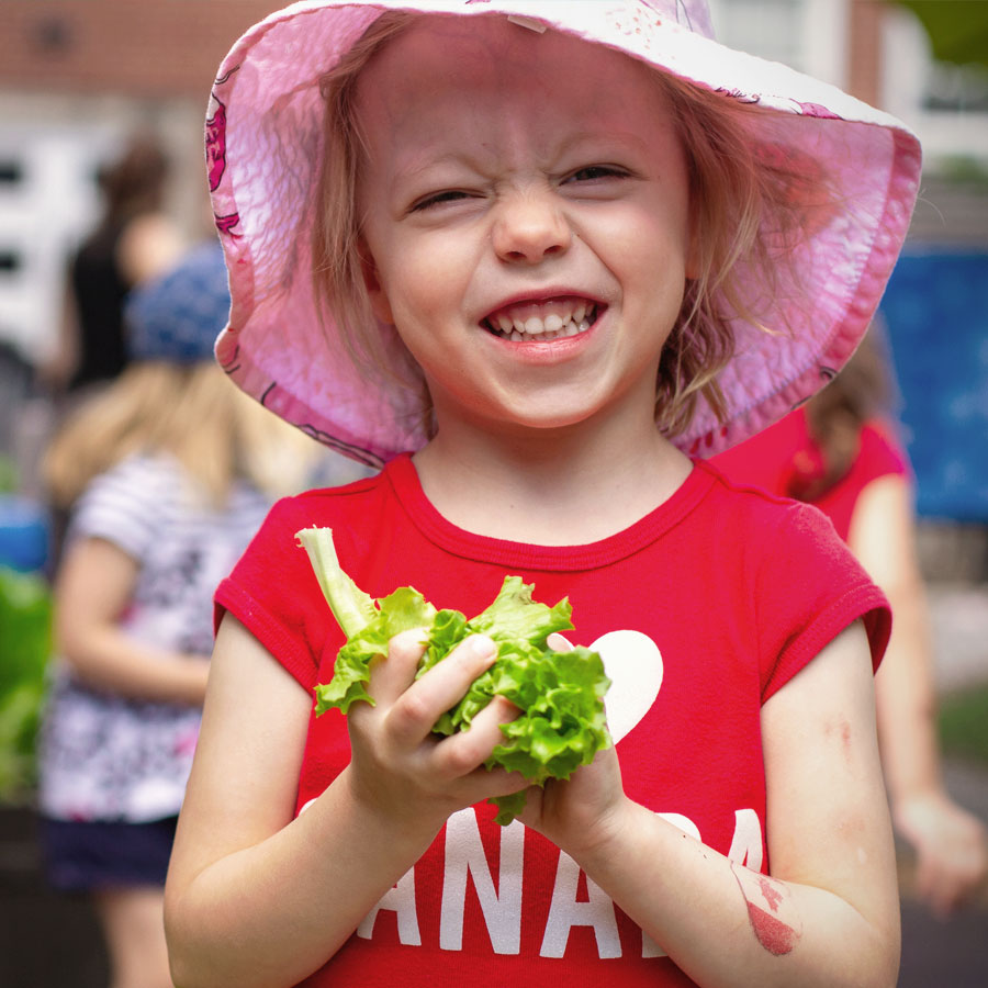 A candid photo of a preschool student at Windermere Kids, smiling and squinting with excitement as she holds freshly picked lettuce from the school's outdoor garden beds. She is wearing a pink sun hat, a red tshirt with a white heart and the word Canada below. A faded temporary Canada tattoo in on her arm.