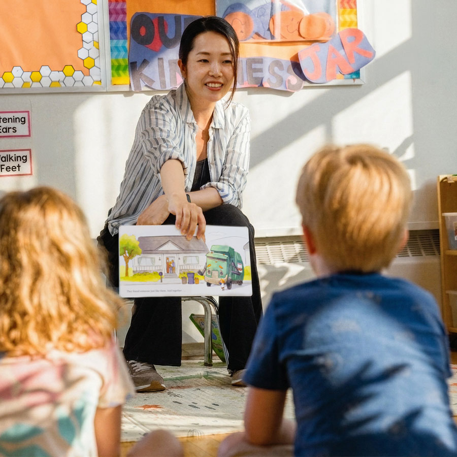 A candid indoor classroom shot of a smiling female ECE teacher showing the illustrations from the book Trash Truck. Two students, one male and one female, are sitting back turned to the camera looking at the illustration. Warm light fills a room with shadows cast from sun entering through nearby windows.