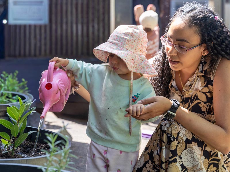 A female ECE teacher holds a toddler's hand as she pours water from a pink watering can into a pot growing lettuce. 