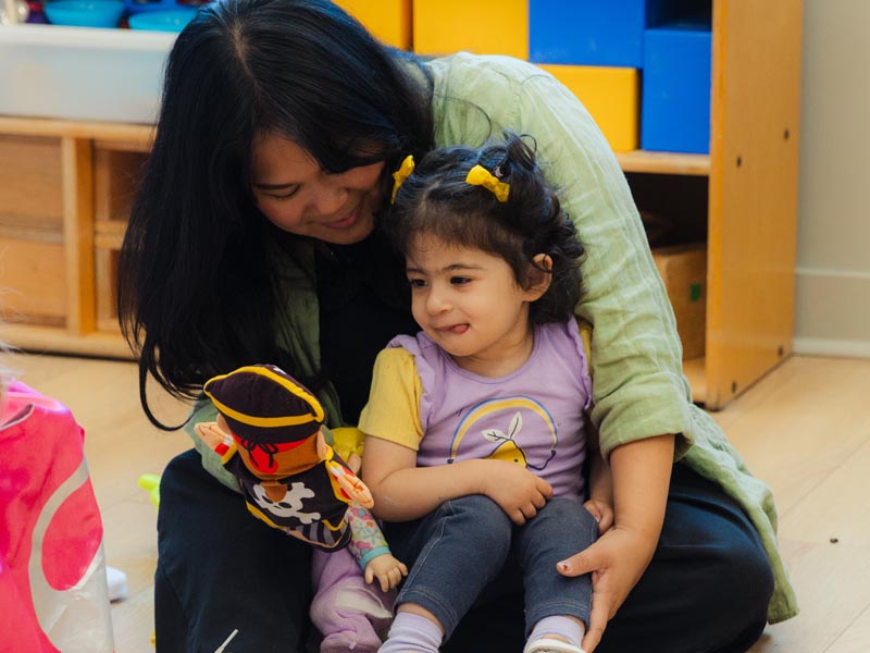 A candid photo of a female ECE teacher holding a toddler in their lap, with a hand puppet on. The child looks secure and happy.