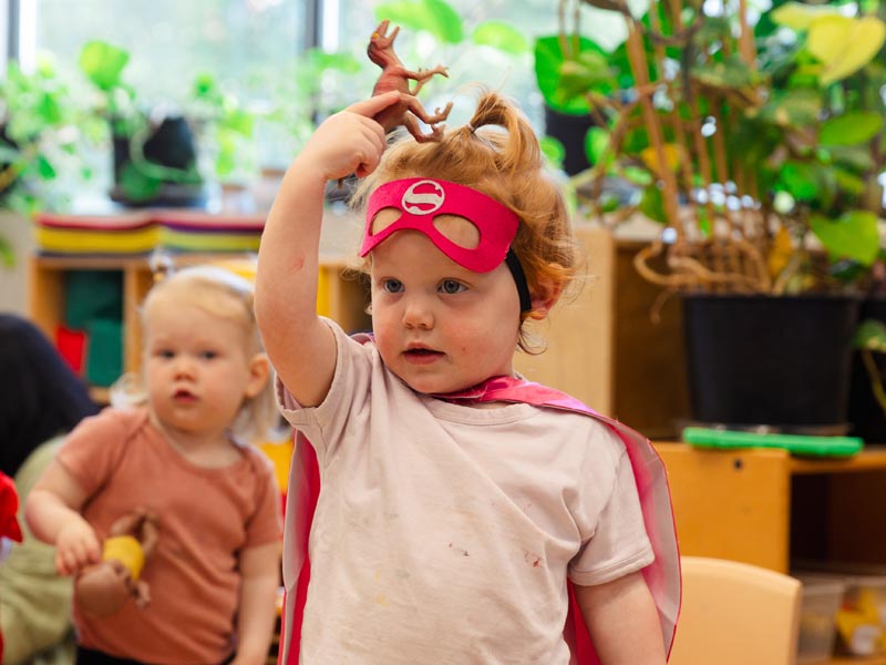 A candid photo of a toddler inside a Green Apple Kids toddler room, holding a toy dinosaur over their head while dressed as a superhero.