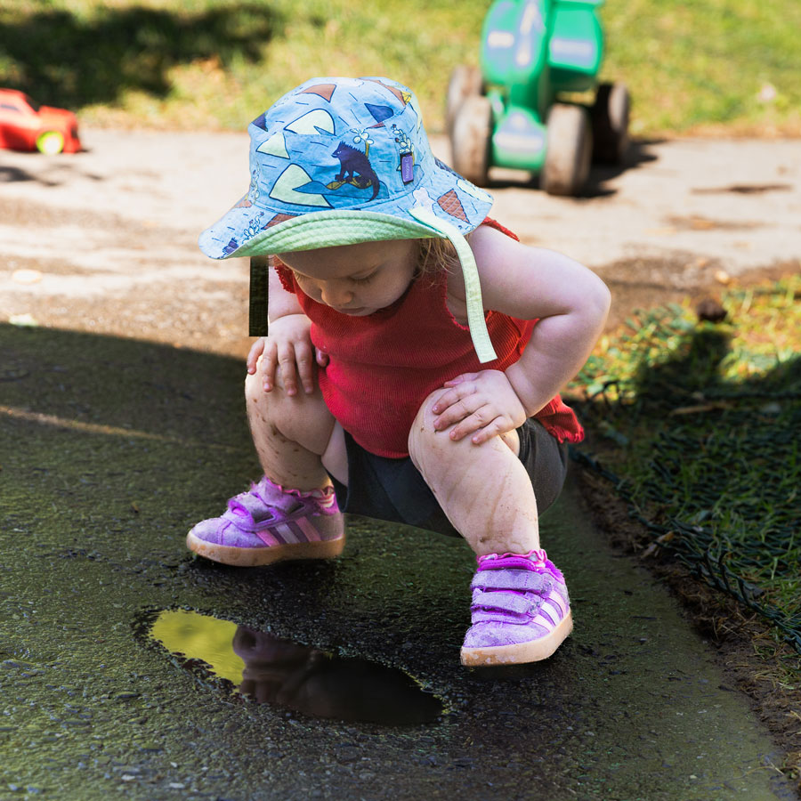 A candid photo of a female toddler kneeling down to study a puddle of water on the asphalt walkway inside Green Apple Kids' natural playground.