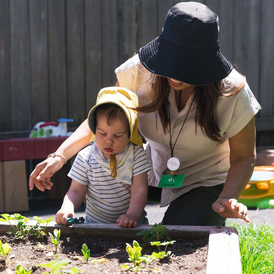 A candid photo of a female ECE teacher kneeling beside an infant who is ready to dig in Windermere Kids' outdoor garden beds. A mix of sunlight and shade illuminate the photo.