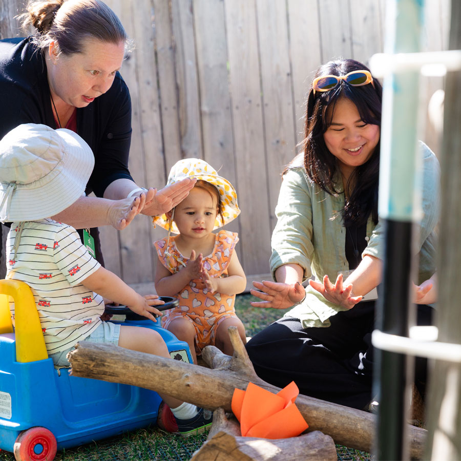 A candid photo of two female ECE teachers siting with 2 toddlers in Green Apple Kids' outdoor playground. They are all imagining they are sitting around a campfire, warming their hands. Real logs are stacked and a piece of orange paper is positioned to simulate the flame from a camp fire.