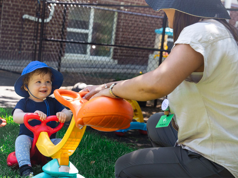 A sunny outdoor candid shot of an infant smiling at the teacher, while the teacher pushes down on a small teeter totter. 