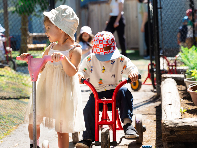 A candid photo of two preschool students riding tricycles on the outdoor track in the natural playgrround at Green Apple Kids. Both children are smiling. Other children and teachers are in the background out of focus.