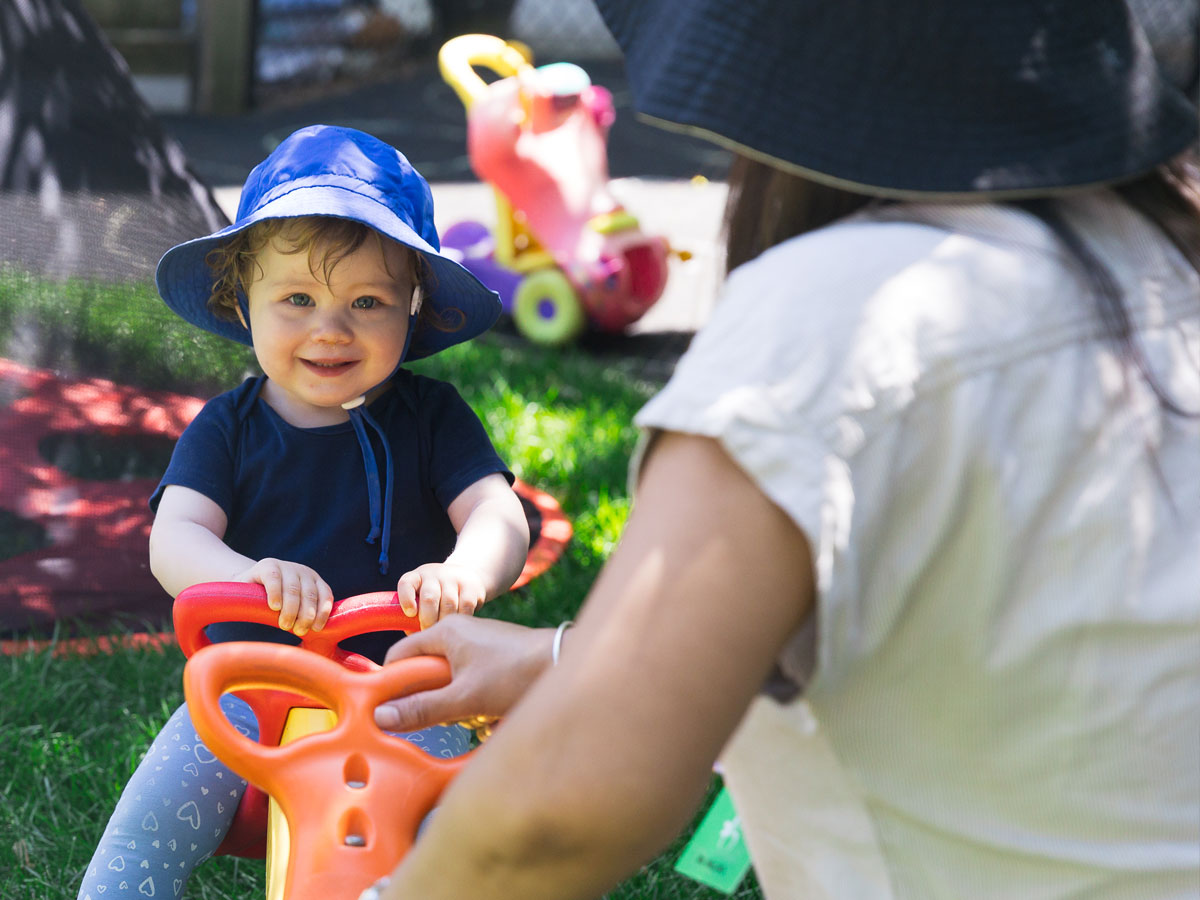 A candid photo of an infant smiling as a teacher pushes down on the teeter tooter the child is sitting on. They are outside in the Windermere Kids playground with a warm natural light illuminating the scene.