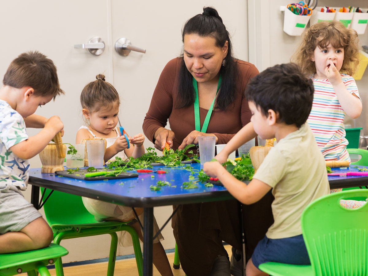 An indoor candid photo of 4 preschoolers sitting at a table with a female ECE teacher. They are focused on cutting and grinding freshly picked parsley from Green Apple Kids' garden beds. Walls are a neutral beige, the table top is blue and chairs are a vibrant green.