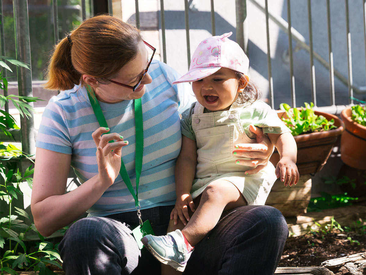 A candid photo of a female ECE teacher balancing a a young toddler child on her lap. The teacher is looking at the student ready to give a playful tickle while the child smiles in anticipation. The Green Apple Kids potted plants and garden bed fill the background with a mix of shade and sunlight illuminating the scene.