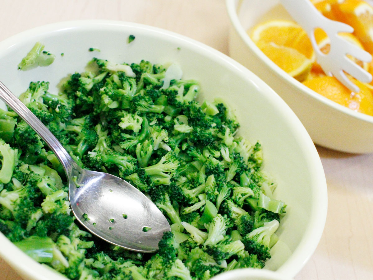A closeup of broccoli with in a bowl with a silver serving spoon as well another bowl of sliced oranges with white serving tongs.