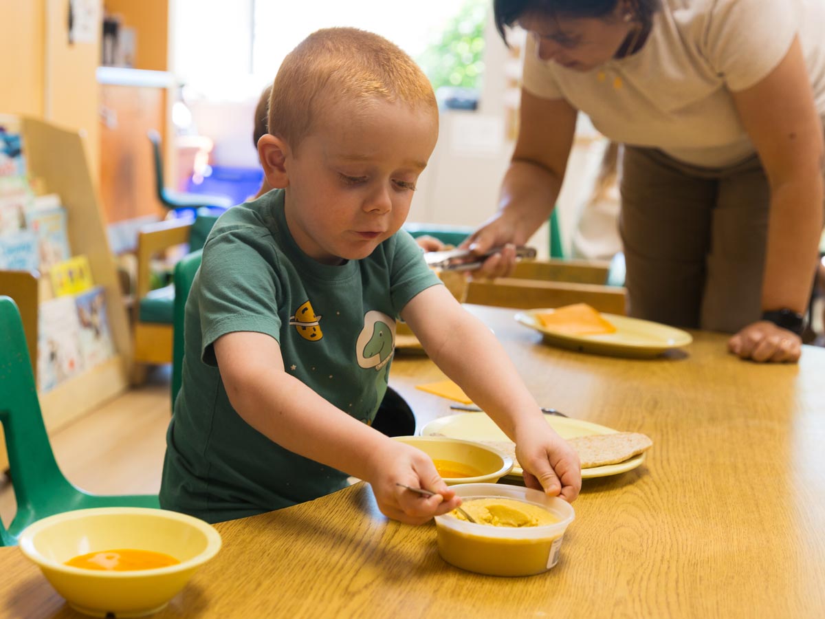 A candid photo of a male preschooler scooping humus from a container while sitting at a desk for lunch. Out of focus in the background is a teacher serving another student.