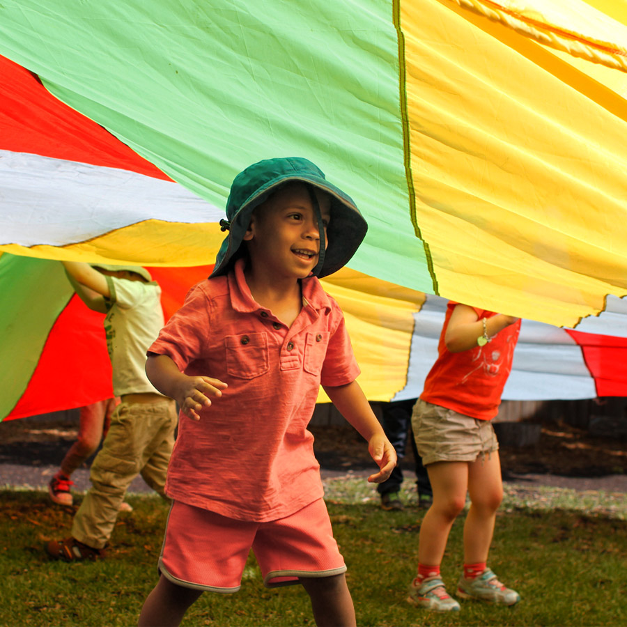 A candid photo of a male Green Apple Kids preschooler smiling underneath a parachute canopy that other children are lifting. The lighting is warm from the parachutes green, yellow, blue and red colours.