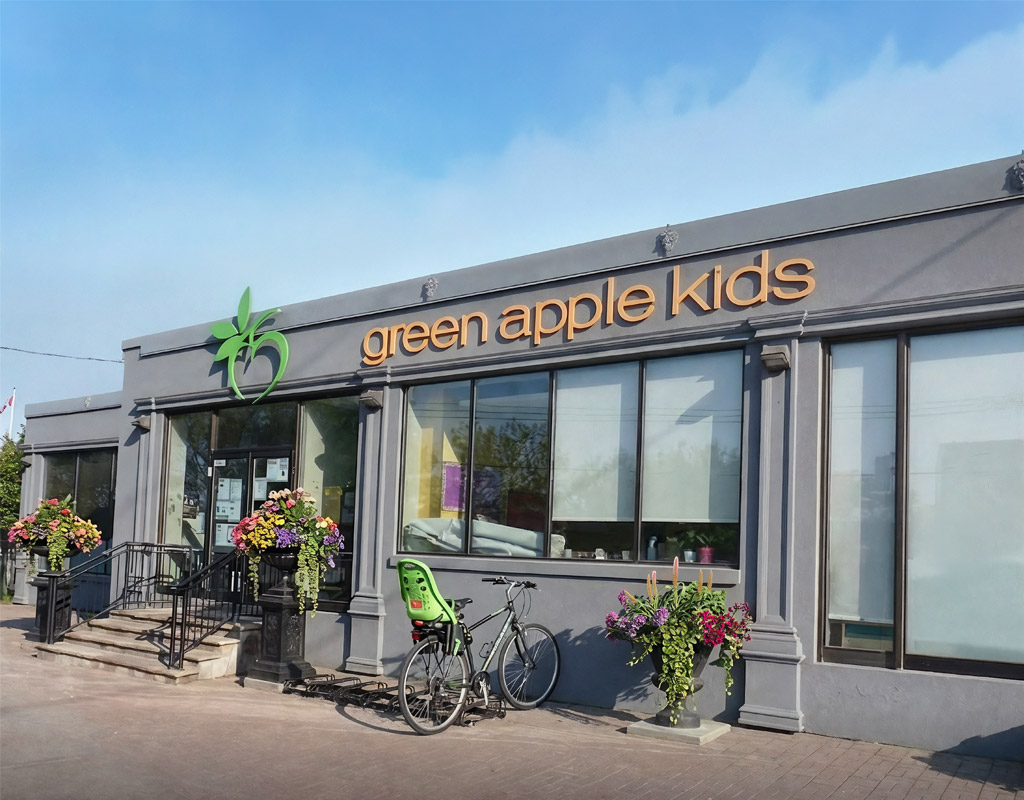 Exterior shot of Green Apple Kids against a blue sky. The building is a modern neutral grey with large windows. Black railings lead to the front entrance with the Green Apple Kids wordmark in Orange and logo in green afixed to the exterior. Outdoor potted plants fill black urns and a bike with a toddler seat on the back is locked to the bike stand.