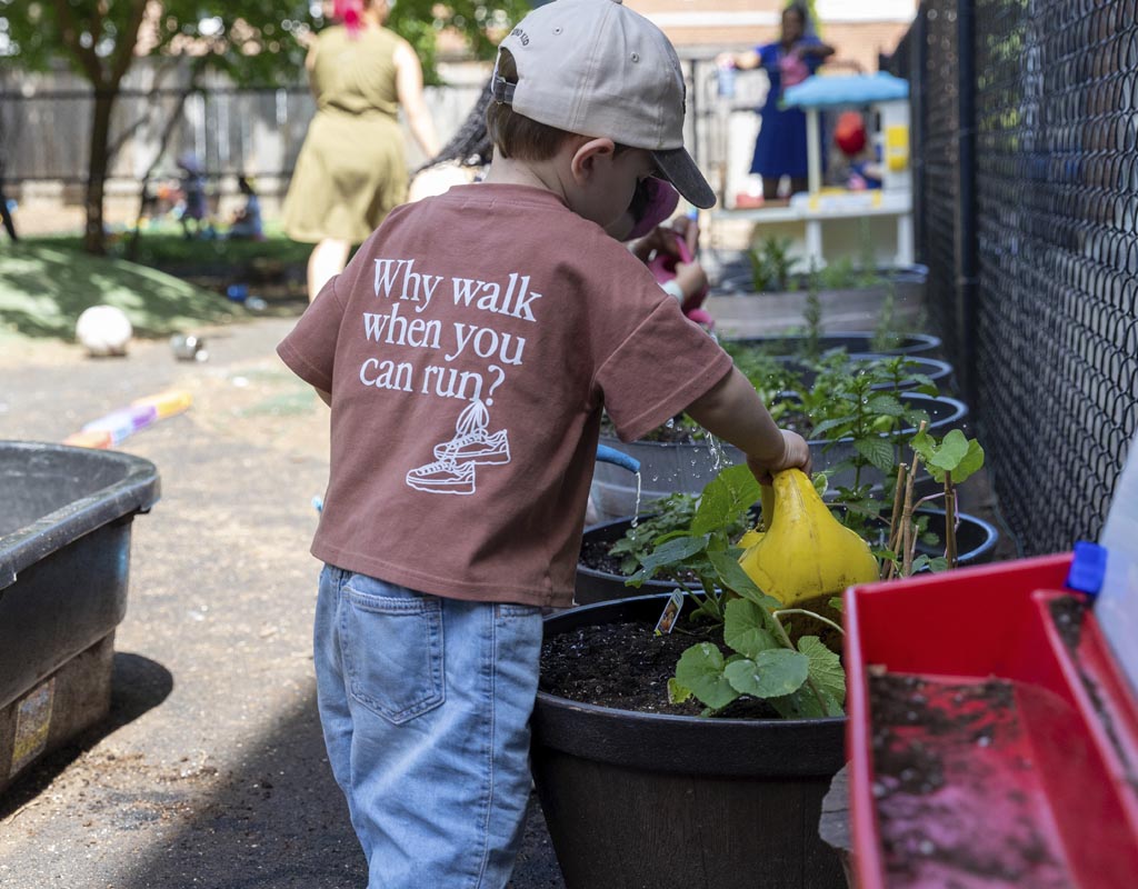 A male preschooler holding a yellow watering can carefully pouring water on potted herbs in WIndermere Kids' natural playground. Two female ECEs can be seen in the background.