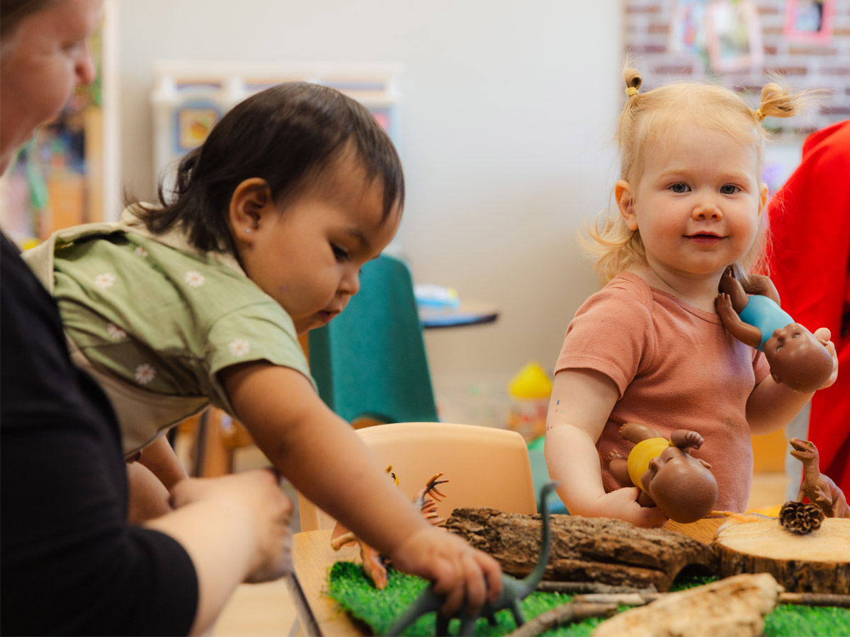 A candid photo of a toddler smiling at the camera holding two baby dolls, one in each hand. Another toddler in the foreground is playing with a toy dinosaur on a table, while being held by a female ECE teacher.