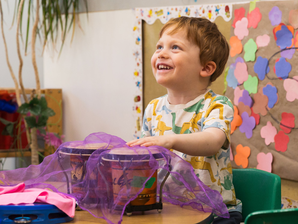 A male preschooler playing on a set of bongo drums while sitting at a desk. He's looking up with a big smile. Student artwork is hung behind him.