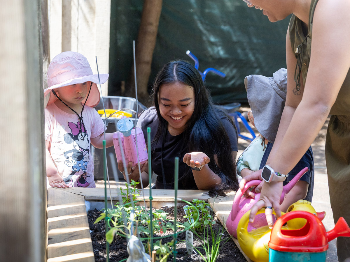 A candid photo of 3 preschools and 2 female ECE teachers huddled around a planter box filled with growing herbs.