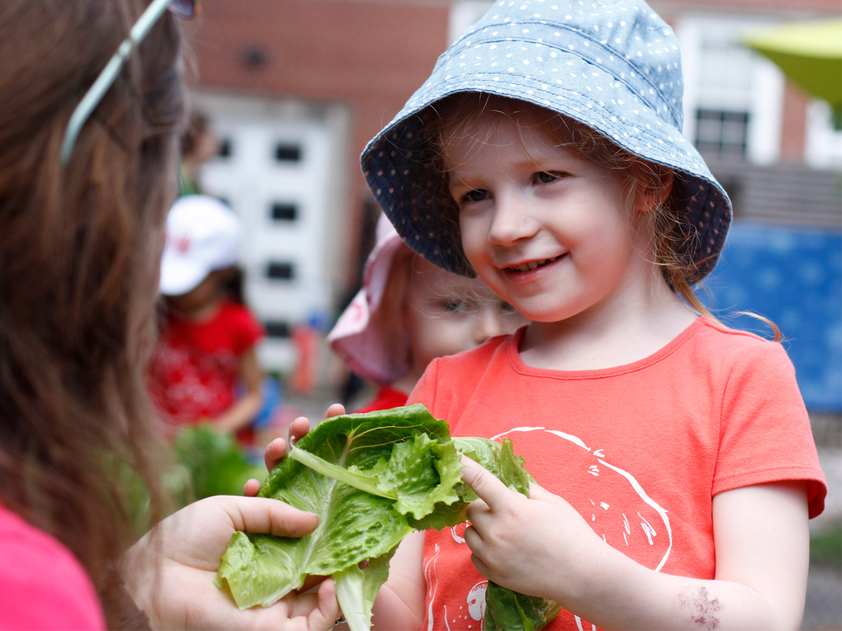 A candid photo of a female preschooler smiling, while passing a few lettuce leaves to a female ECE.